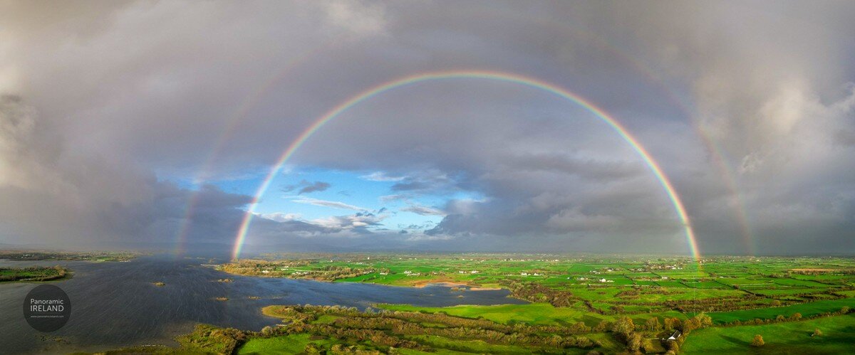 Image: A sweeping panoramic, aerial view of a spectacular double rainbow arching over a vast Irish landscape. The primary rainbow is bright and forms a complete, perfect arc, with a fainter secondary rainbow visible above it. Beneath the rainbows, the land is a vibrant patchwork of bright green fields, dotted with small houses, trees, and stone walls. To the left, a large, dark body of water, likely a lough or wide river, reflects the sky. The weather is dramatic: dark, heavy storm clouds frame the scene on both sides, while a patch of bright blue sky with white clouds shines through directly under the rainbow's peak, indicating the sun is breaking through. Image by Panoramic Ireland.
