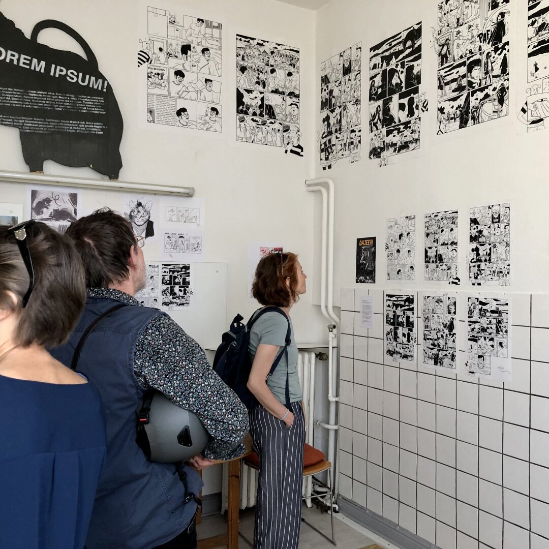 Menschen stehen in einem gekachelten Raum und schauen sich eine Comicausstellung an der Wand an. Gezeigt wird mein Beitrag zu einem Fanzine, in dem ich verschiedene Comicfiguren aufgreife, die mich als Kind begleitet haben. Es ist ein Comic in Schwarzweiß.

Oben im Bild steht auf einer Neonröhre eine schwarze Ratte aus Kappa-Platte, auf der in Weiß ein Fülltext aufgesprayed ist.