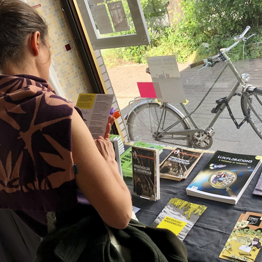 Ein Mensch schaut sich am Büchertisch einen Flyer an. Es liegen Bücher, Hefte und Flyer mit Arbeiten von mir auf dem Tisch.