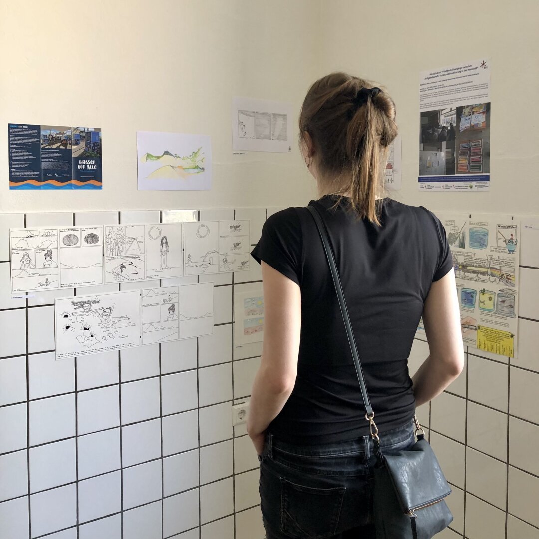 Ein Mensch steht vor einer gekachelten Wand und schaut sich Arbeiten von Teilnehmer:innen eine Comic Workshops zum Thema Wasser an.