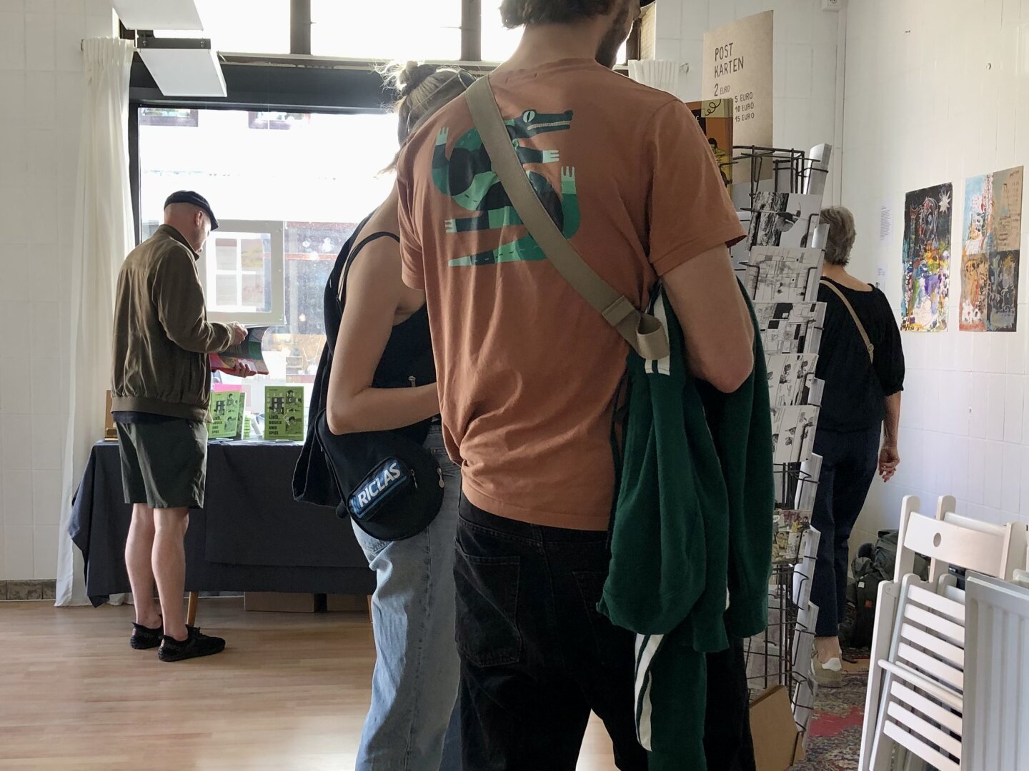 Blick in den Raum. Menschen schauen sich Postkarten am Postkartenständer und Bücher vom Büchertisch an.