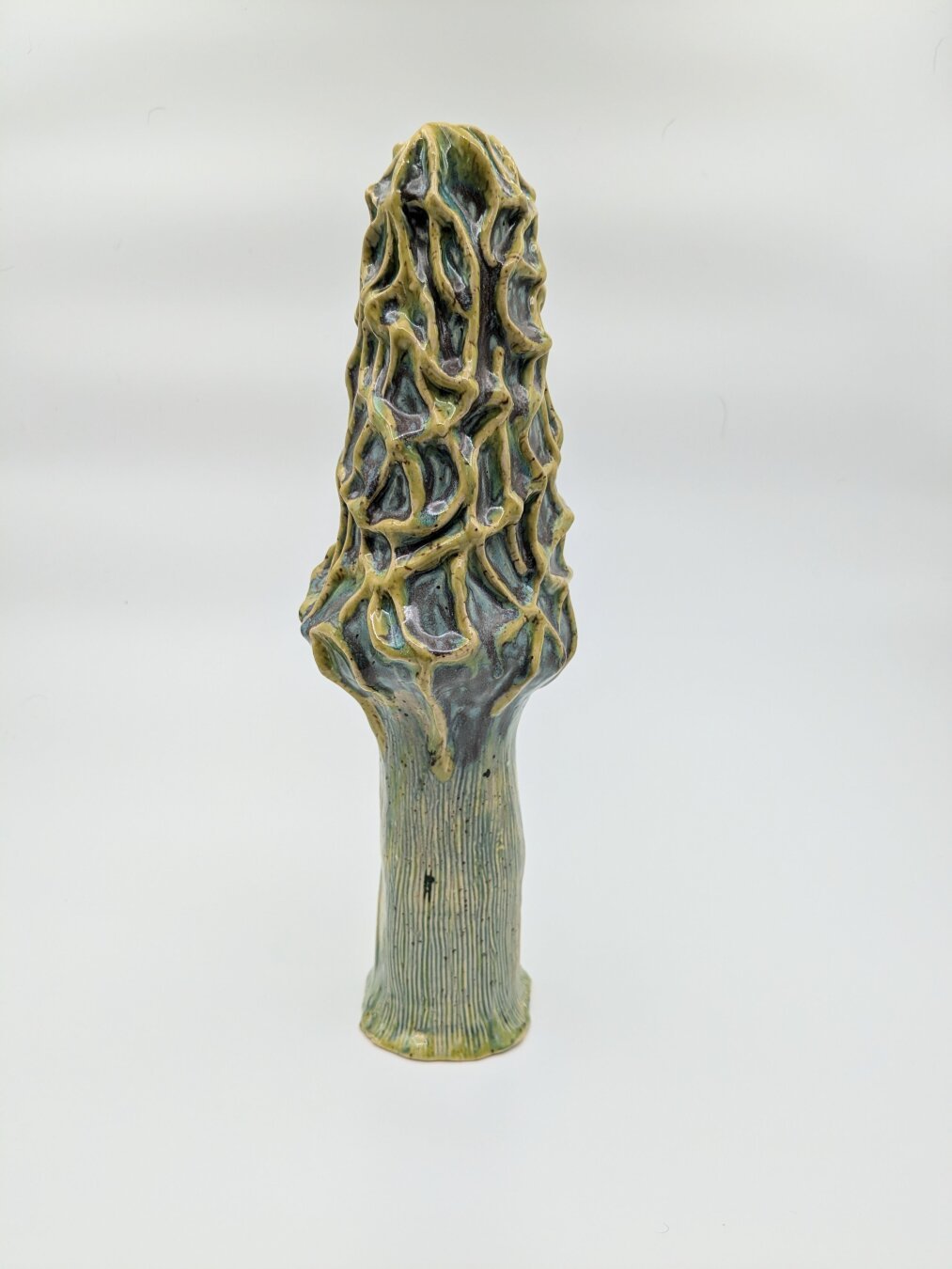 A ceramic statue of a morel mushroom in bright colors not typical of a real morel mushroom. A chartreuse green is used for the body of the mushroom, creavaces have a muddy purple/ blue applied to enhance the depth of the surface textures.