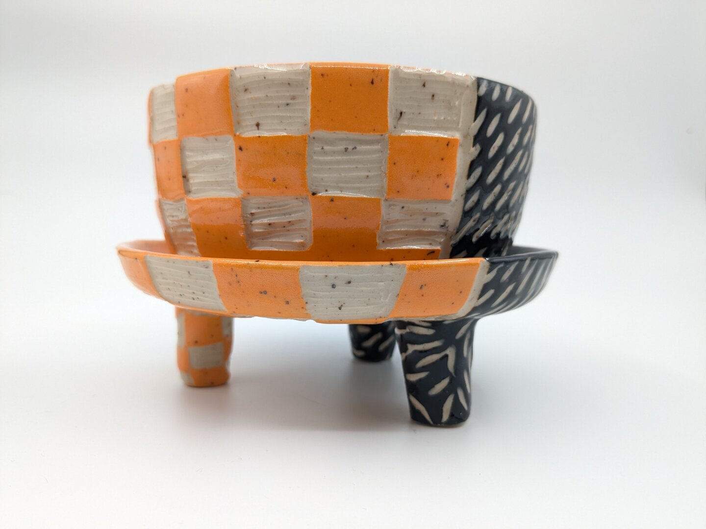 A handmade planter with attached drainage and mid century modern style feet. The planter is half orange and half black. The orange side of the planter has a checkered pattern carved into the surface of the clay, revealing the clay body underneath. The black half of the planter has dashes carved diagonally into the surface of the clay - the carved dashes show the speckled white clay body underneath.