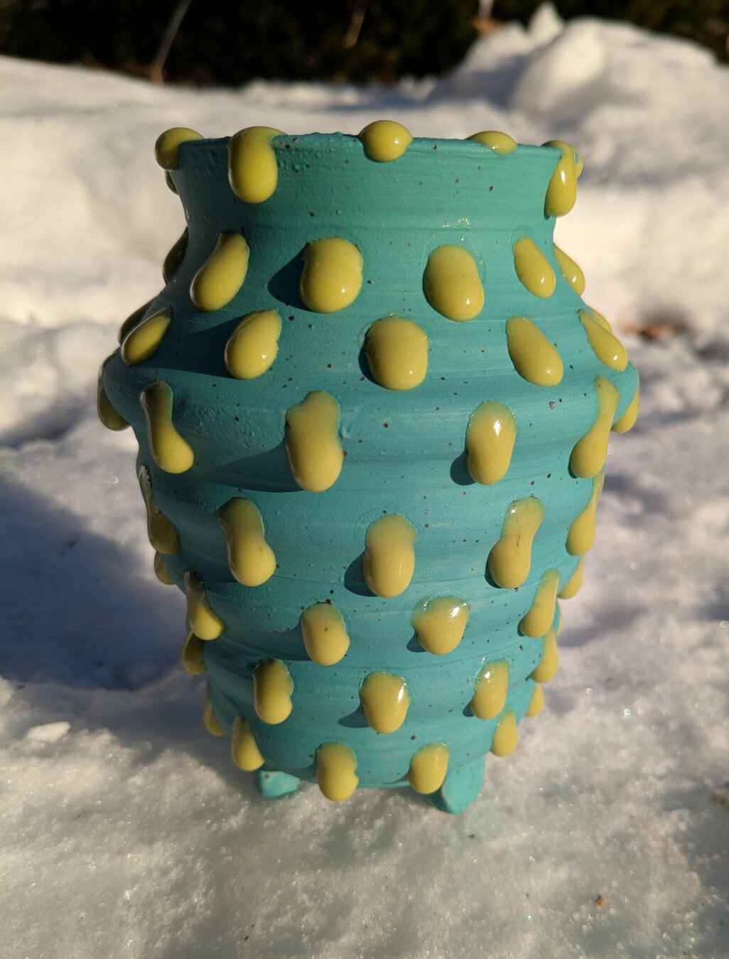 A marine blue stoneware vase with small block feet. The vase walls are dotted with shiny blobs/droplets of ooze green glaze all along the exterior of the vase