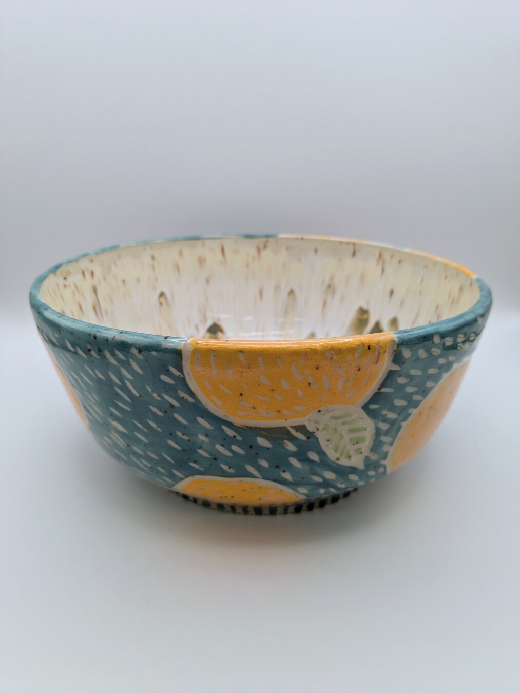 Handmade wheel thrown fruit bowl. The bowl is colored turquoise blue and has oranges randomly patterning the exterior of the bowl. Lines have been carved into the bowl to define the shape of the oranges and leaves. The interior of the bowl is slightly visible showing a light tan color with streaks of darker tans and browns.