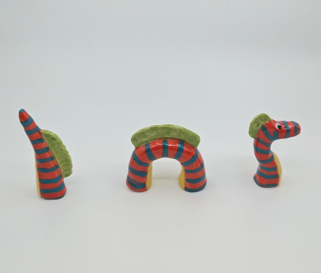 A three piece miniature ceramic sculpture of a serpentine-like dragon, consisting of a head, middle, and tail pieces on a white background. It is brightly colored, the body is orangey-red with turquoise blue stripes running horizontally across the dragon body, the underbelly is a sunny yellow and the dorsal fins are lime green.