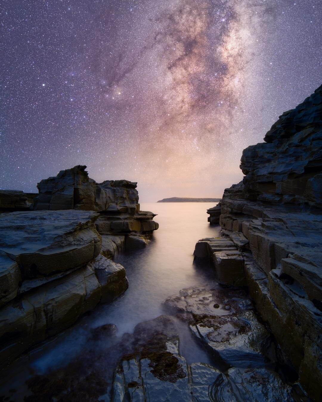 The Milky Way dominates the night sky above the rugged basalt cliffs of Cape Woolamai on Phillip Island. Smooth, long-exposure water flows through a narrow rocky inlet toward a distant headland, while countless stars and intricate galactic dust clouds fill the sky above the dramatic coastal landscape.