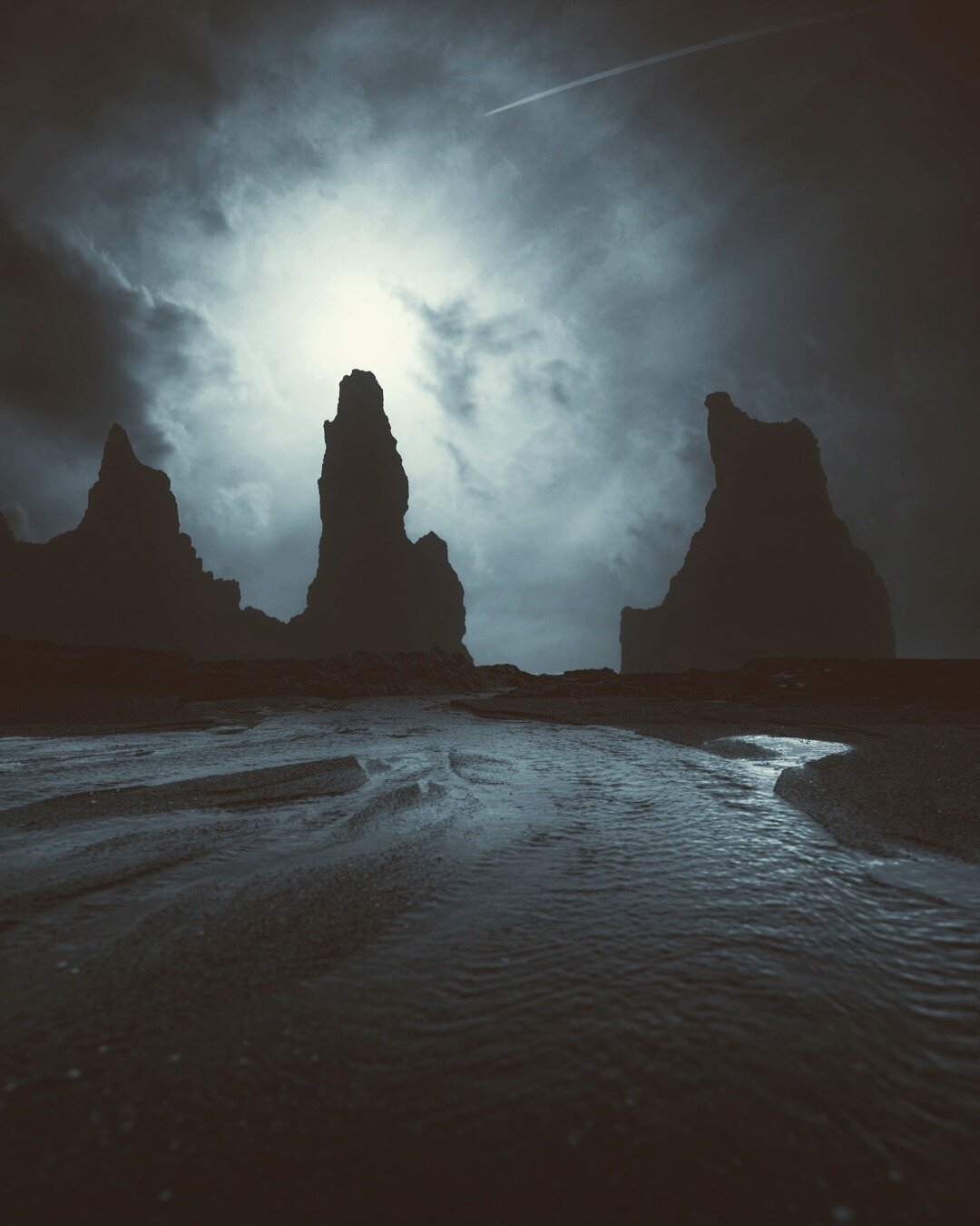 A full moon shines through heavy clouds behind towering Icelandic sea stacks, casting a cold glow across a dark volcanic beach with shallow water flowing through the foreground.