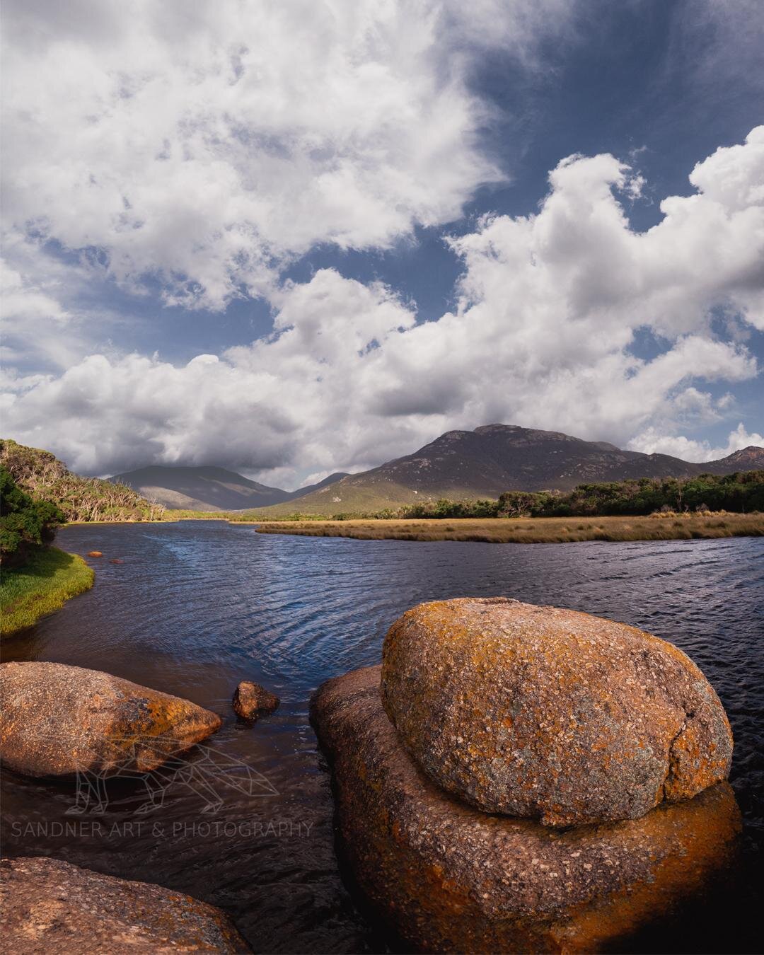 Granite boulders frame a dark river at Wilsons Promontory National Park beneath dramatic white cumulus clouds and distant granite mountains, illuminated by bright afternoon sunlight.