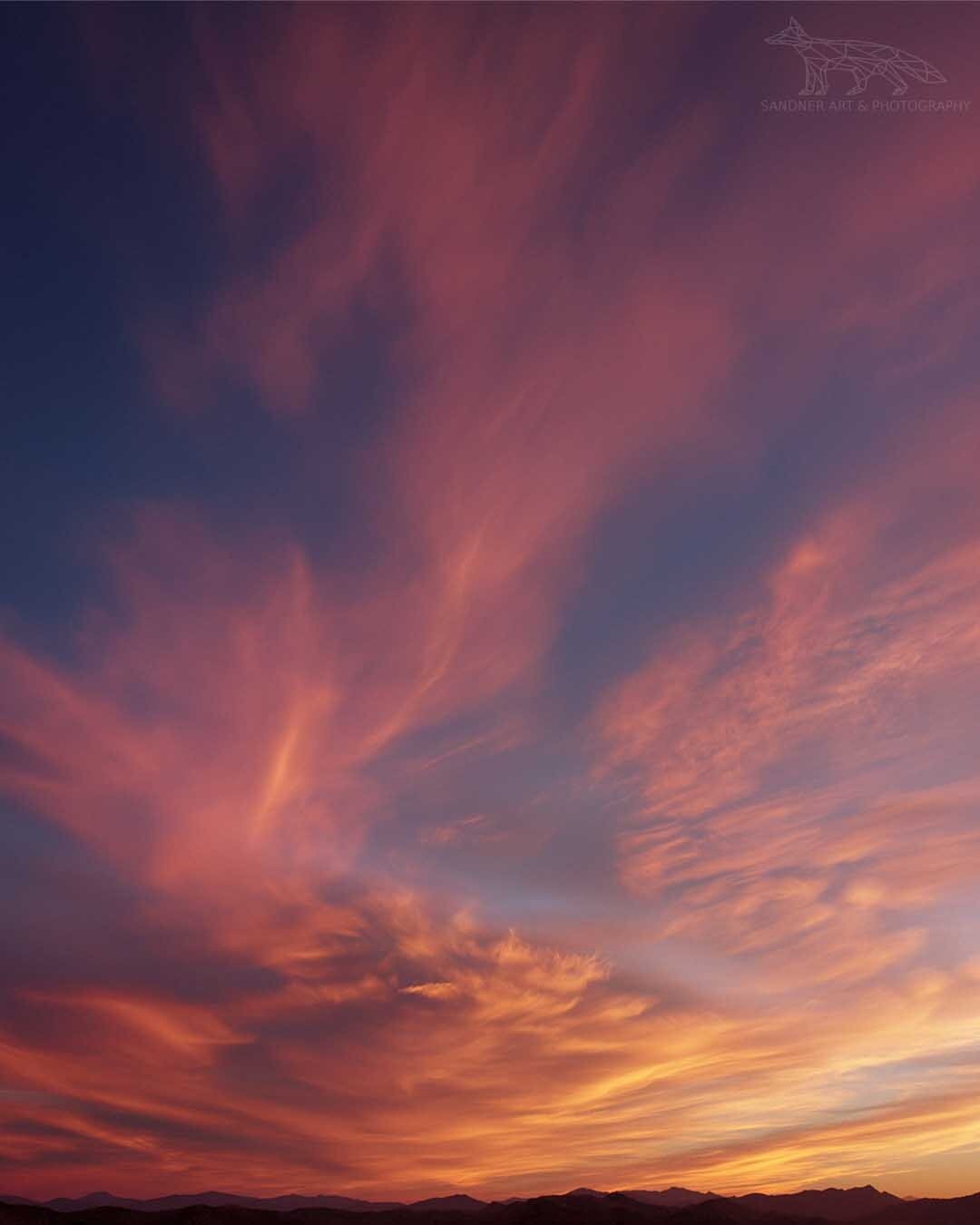 A vibrant New Zealand sunset fills the sky with sweeping shades of pink, orange, and purple. Delicate wisps of cloud stretch across the horizon above a distant mountain range, while the last warm light of the day glows beneath the colourful evening sky.