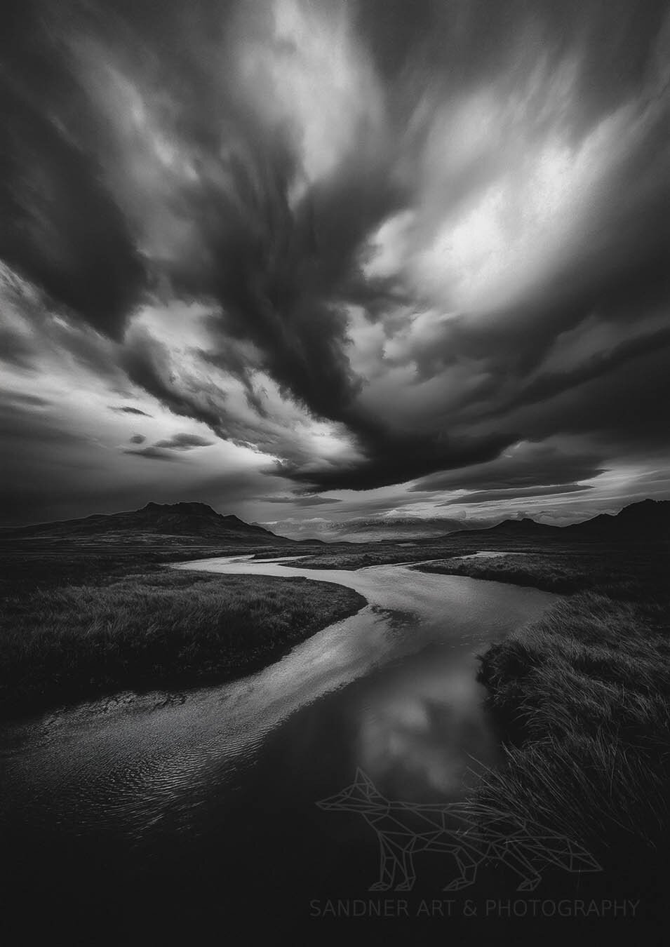 A dramatic black-and-white landscape featuring a winding river cutting through an open plain, leading toward distant mountains. Above, dark storm clouds swirl with intense contrast, creating a moody and atmospheric scene. The river reflects the sky, adding depth to the composition.