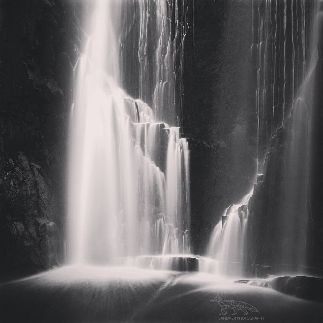A black-and-white long-exposure photograph of Mackenzie Falls in the Grampians National Park, Victoria. Water cascades in soft, silky streams down a dark cliff face, pooling into a misty basin below. The contrast between the bright flowing water and the shadowy rock walls creates a dramatic, timeless scene.
