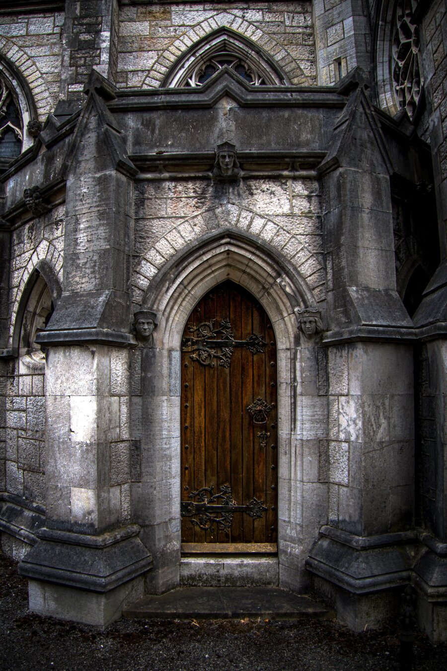 A close-up of a weathered, dark brown oak door with vertical wood planks, set in a pointed Gothic grey stone archway. The door is adorned with intricate, black wrought-iron hardware, including two large decorative vine shaped strap hinges on the left and a ring latch on the right. Carved stone figureheads frame the doorway on the left, right, and top of the arch, surrounded by aged, textured stone masonry.