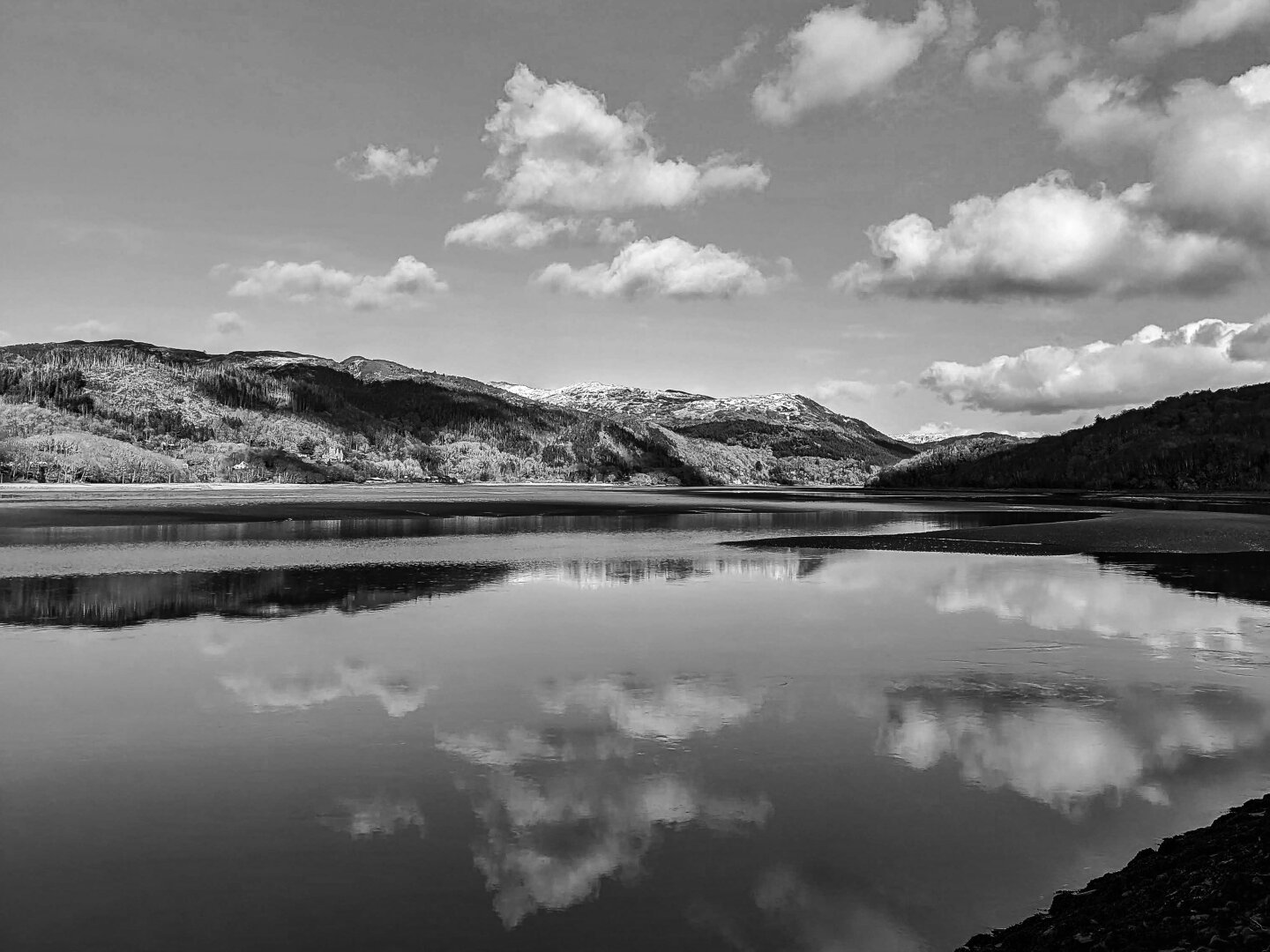 A black and white landscape of a calm, wide estuary reflecting a cloudy sky and distant mountains. Rolling hills stretch across the horizon, with light patches of snow visible on the highest peaks. The smooth water creates a mirror image of the fluffy white clouds above.