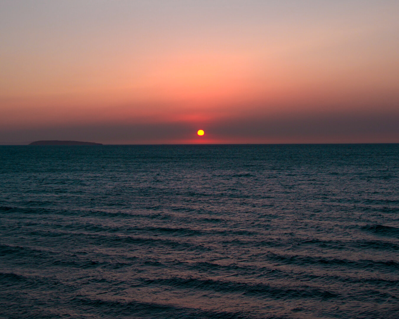 A vibrant orange sunset over a calm, dark blue ocean with a small island silhouette on the left horizon. A small, bright orange sun sits just over a dark horizon line under a sky shifting from pink to orange. Gentle, dark ocean waves take up the foreground.
