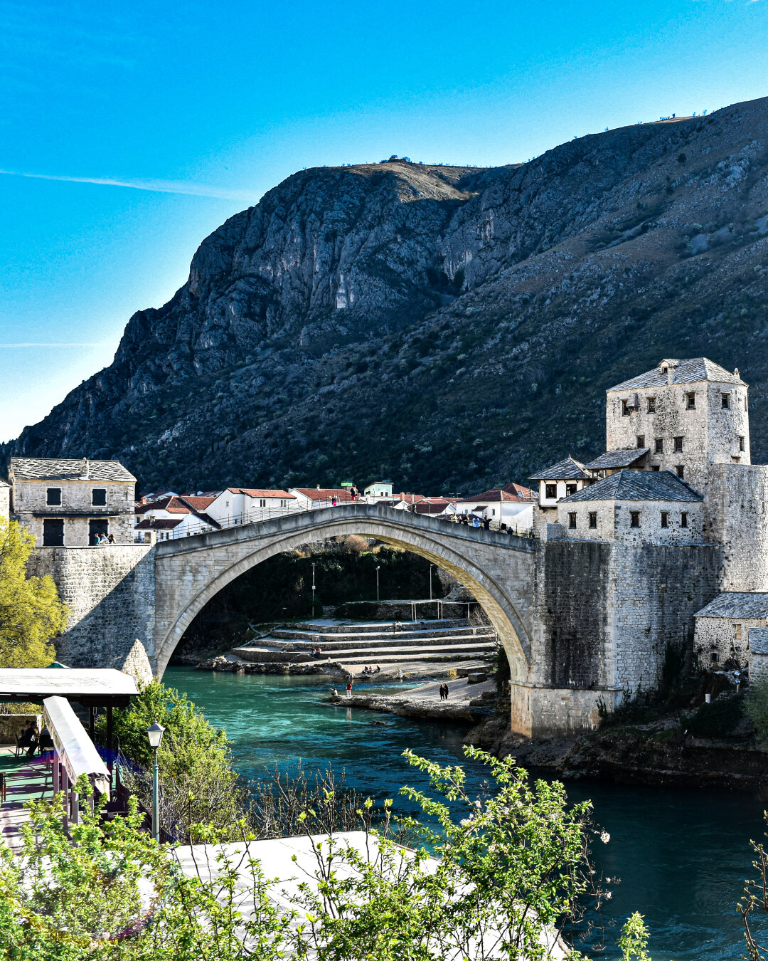 The Stari Most stone arch bridge crossing over the turquoise green Neretva River in Mostar, Bosnia and Herzegovina with historic stone towers at each side. In the background, a steep, rocky mountain rises up to a clear blue sky.