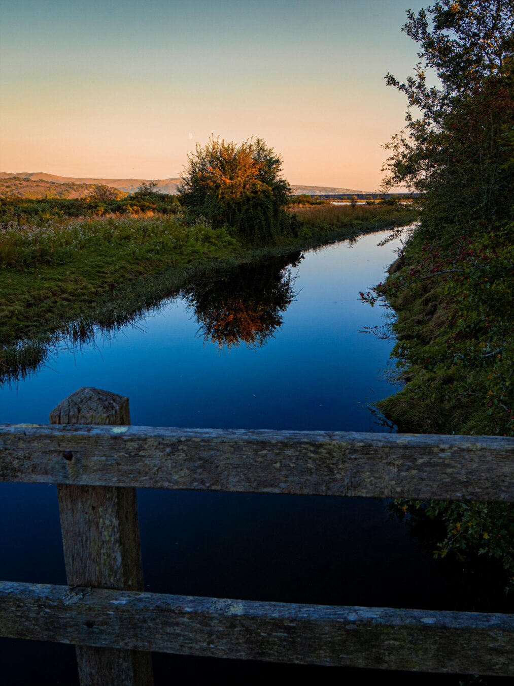 A calm waterway viewed from a rustic, weathered wooden bridge. The still water creates a mirror reflection of a lone bush on the bank and a blue and orange sky. In the background, rolling hills sit under a sunset gradient of soft orange, peach, and blue.