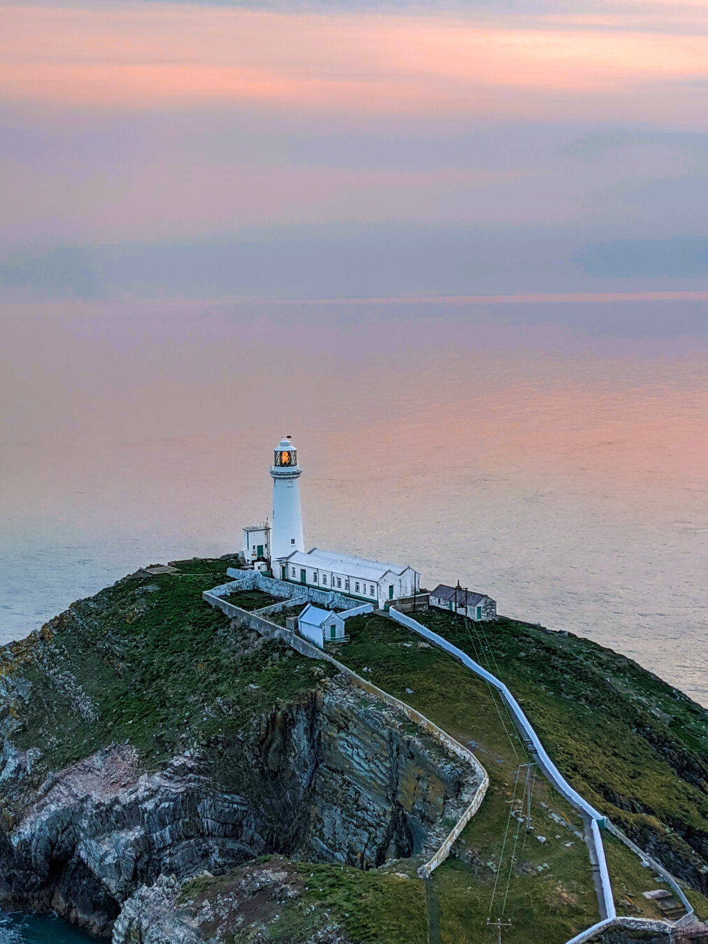 High-angle view of a white lighthouse and its outbuildings at sunset. The lighthouse, with its beacon glowing orange reflecting the sun, stands on a small islet at the bottom of a steep, rocky cliffside in North Wales. A white walled path winds down the green hill. In the background, the pastel pinks of sunset reflect on the water so that you can barely tell where one begins and the other ends.