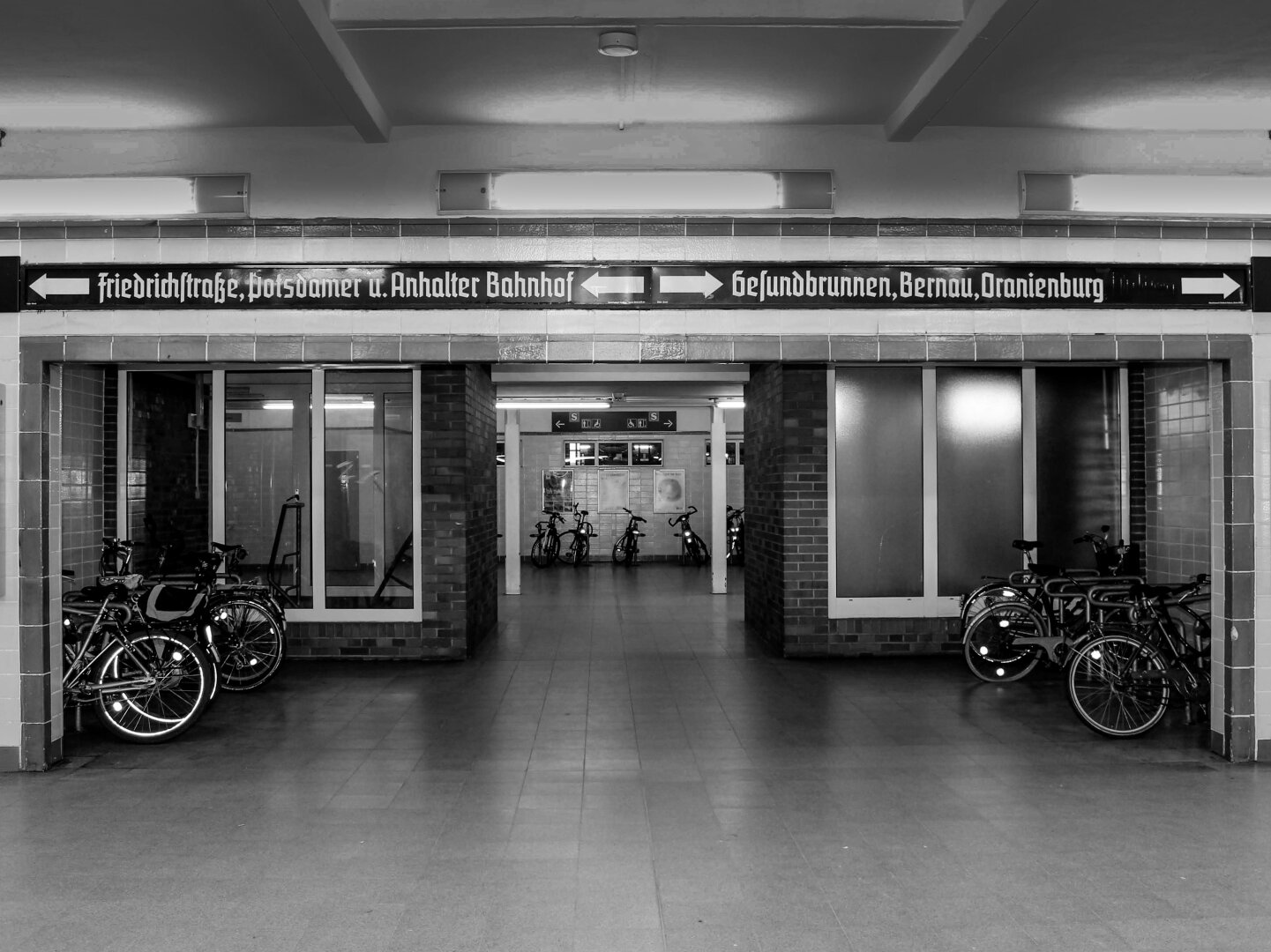 Monochrome photo of an underground station in Berlin. Above a wide rectangular opening, a long, dark sign features white Gothic-style text and arrows pointing left to "Friedrichstraße, Potsdamer u. Anhalter Bahnhof" and right to "Gesundbrunnen, Bernau, Oranienburg." The passage in the centre is filled with bicycles parked in racks on both sides and more parked through to the back wall.