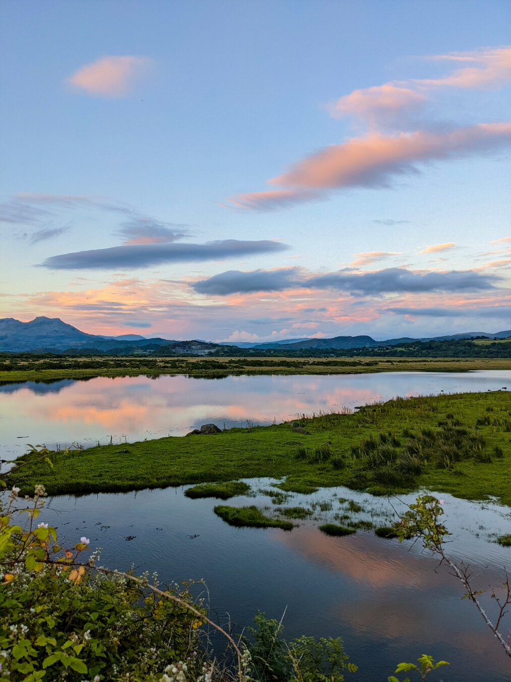 A tranquil landscape overlooking an estuary in North Wales at sunset. In the foreground,  the water reflects the pastel sky with a lush green grassy bank cutting through it. In the background, the distant mountains of Eryri (Snowdonia) rise under soft pink and purple clouds in a light blue sky.