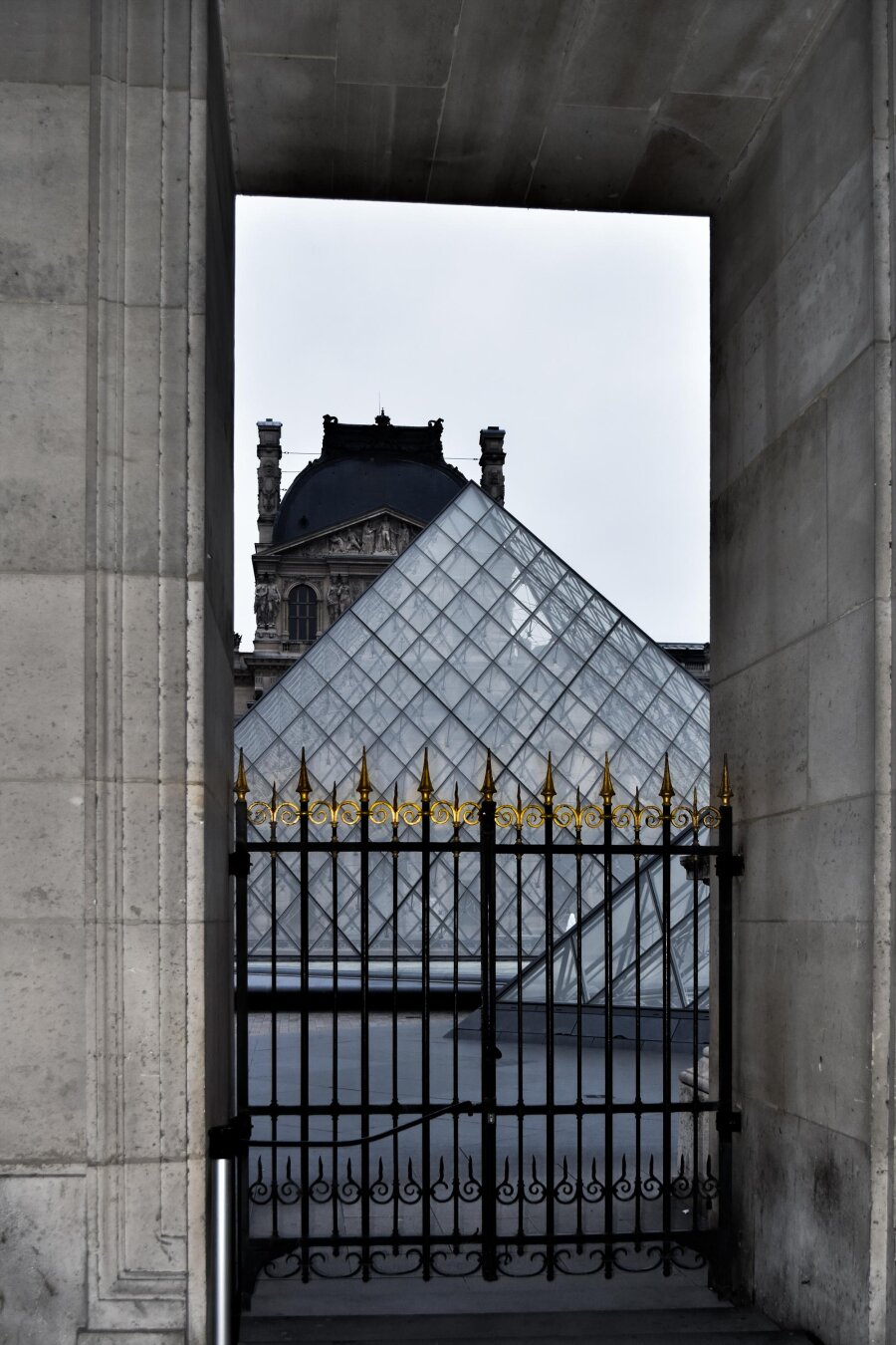 The Louvre Pyramid as seen through a huge stone archway. In the foreground, a black iron gate with ornate gold-painted spikes stands before the glass and steel pyramid. In the background, behind the pyramid, the Renaissance-style roofline of the Louvre Palace can be seen under a white, overcast sky. The image is mostly monochrome but for the golden spikes on the gate.