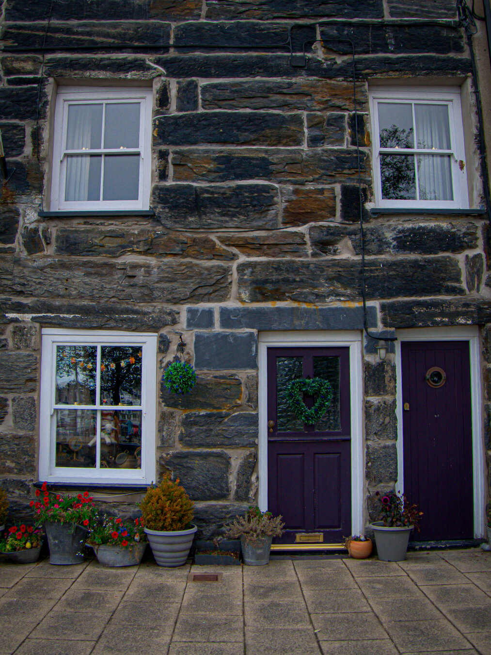 A traditional stone cottage in North Wales. The facade is made up of large, dark Welsh slate blocks with white sash windows. On the ground floor two adjacent deep purple doors sit on the right next to a big white sash window on the left. The main door is a split stable-style with a green heart-shaped wreath hanging over the window panes in the top half. The smaller one is a narrow, solid door made of vertical slats with a small brass portal toward the top. Bright red, blue, and yellow potted flowers, along with miniature shrubs, line the stone patio pavement beneath the window.