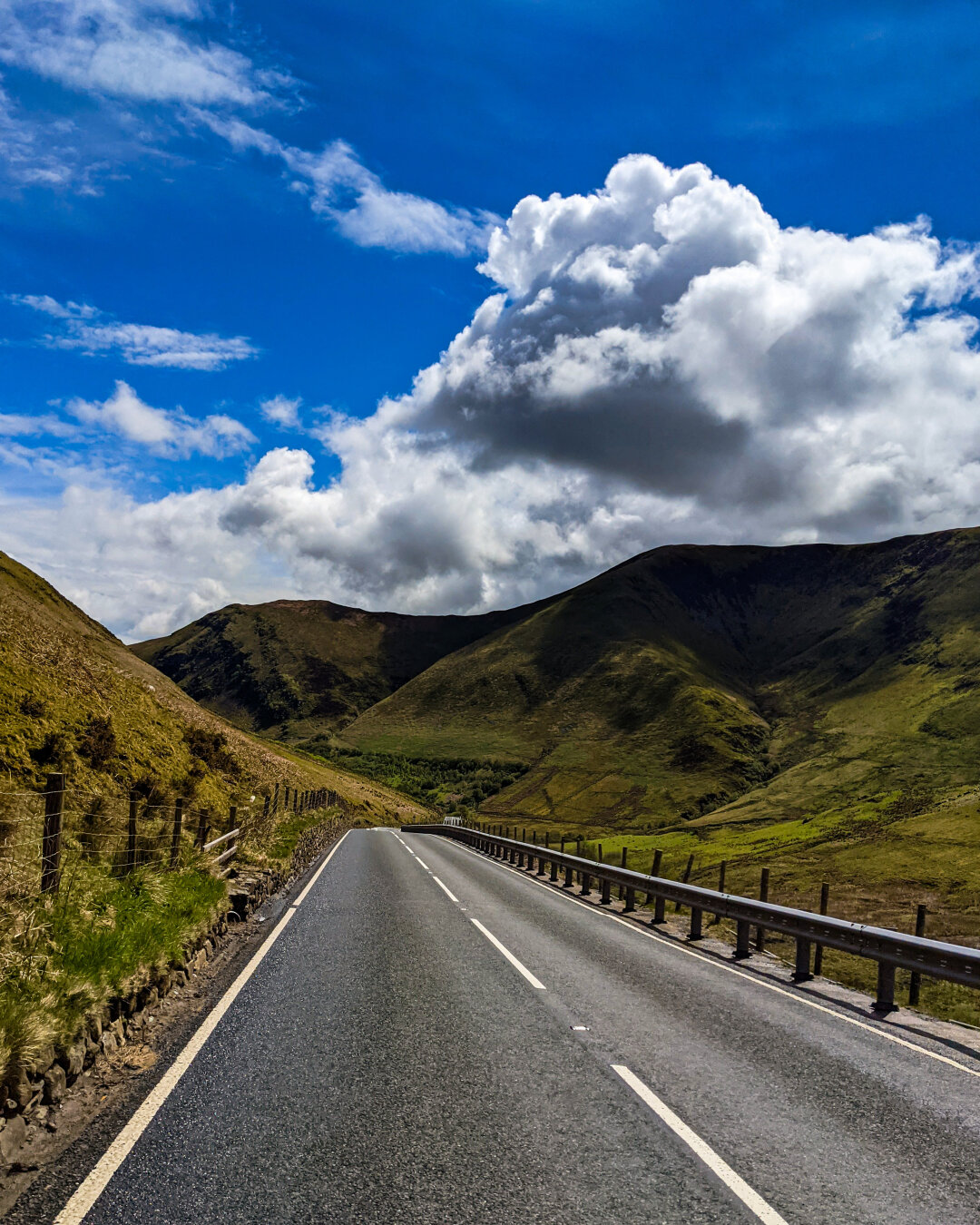 An empty two-lane road curves gently through a valley in Eryri (Snowdonia) on a bright sunny day. Steep, grassy green hills flank the road, and mountains rise ahead under a vibrant blue sky filled with massive, billowing white clouds.
