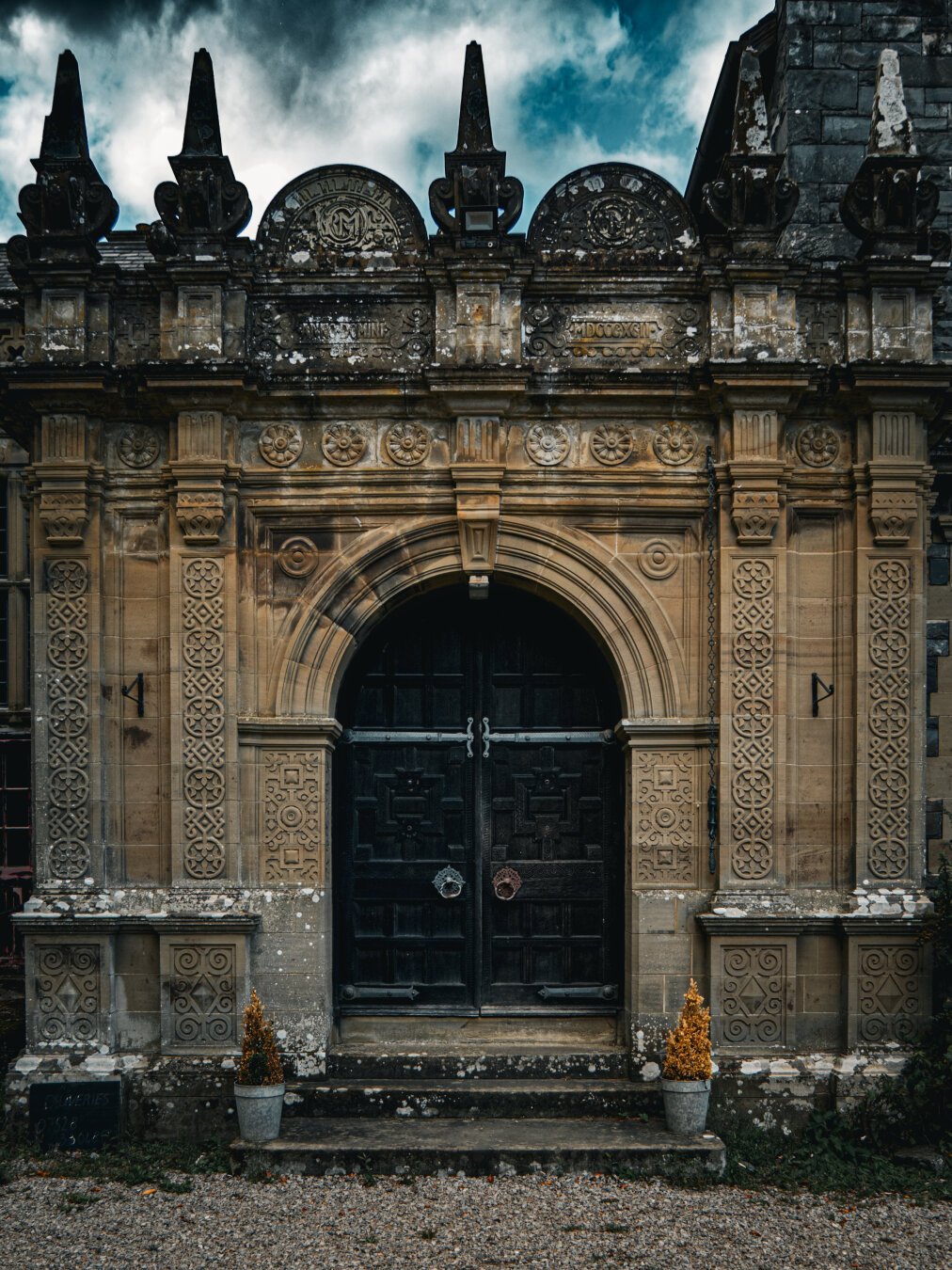An ornate stone entrance porch with heavy arched black wooden double doors featuring raised geometric paneling and large metallic ring knockers. There are intricate geometric carvings on the sandstone pilasters and over the door. At the top of the porch, pointed finials rise into a moody sky.