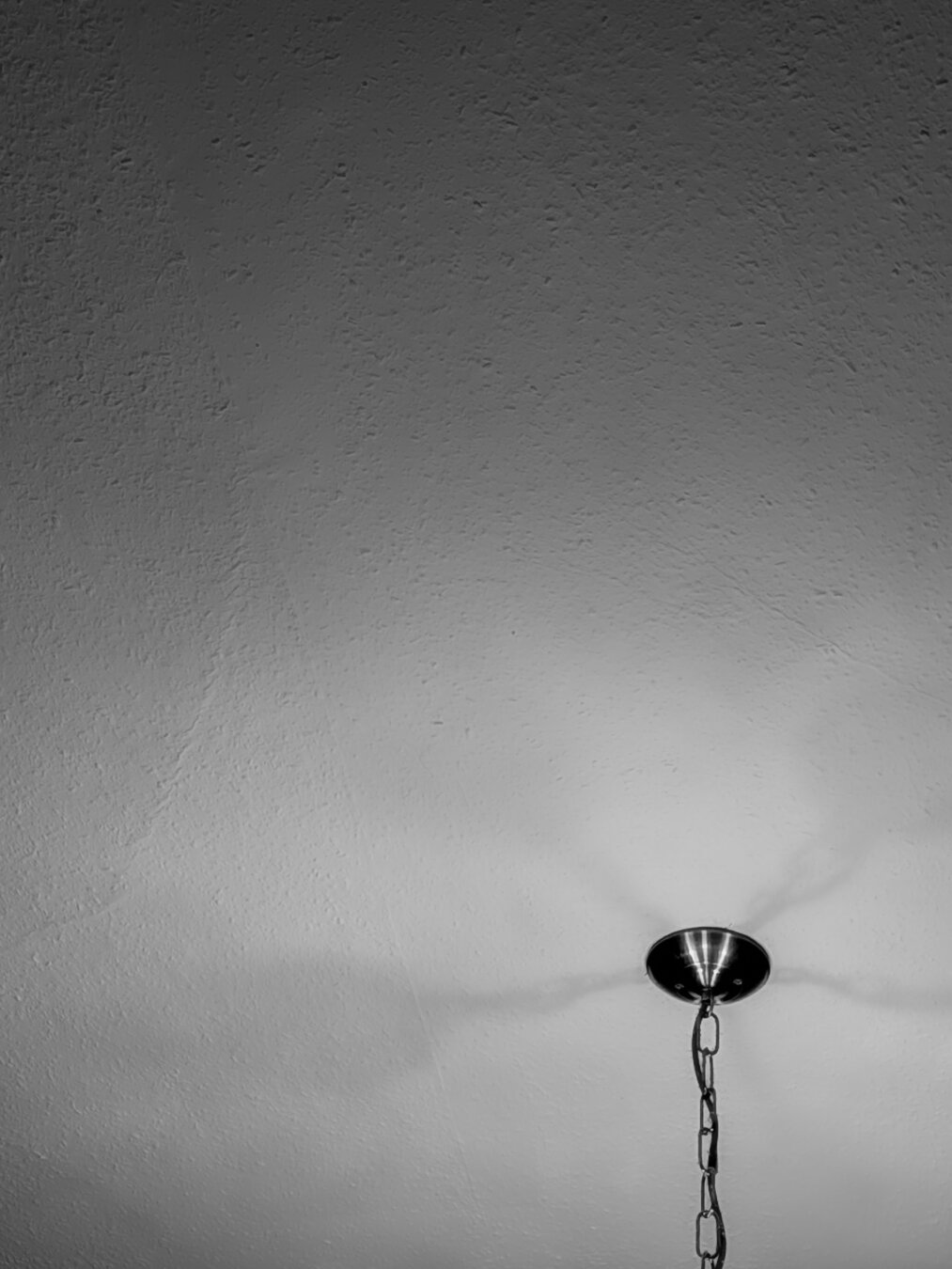 A high-contrast, monochrome photograph looking straight up at a textured plaster ceiling. A dark metal light fixture chain hangs from the ceiling in the lower right corner of the image. The unseen lamp casts shadows against the uneven ceiling.