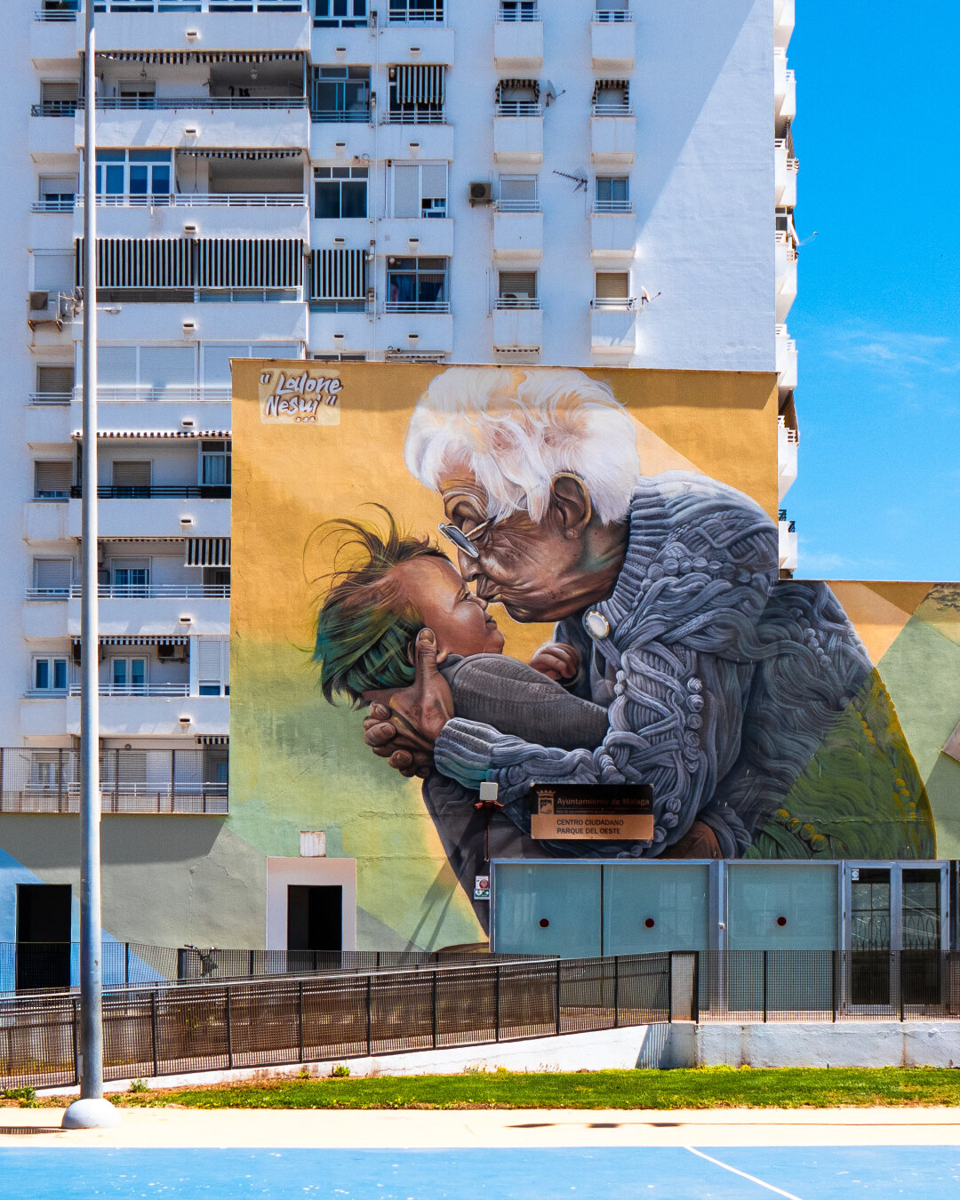 A sunny shot of a large scale, hyper-realistic mural painted on a building. The mural depicts a white-haired elderly woman with glasses holding very young child. The woman wears a gray knitted cardigan and her lips are pursed as she kisses the smiling child on the nose. In the upper left corner of the mural is the text "Lalone Nesui...". A tall modern white apartment building rises in the background under a blue sky and a sports court is visible in the foreground.