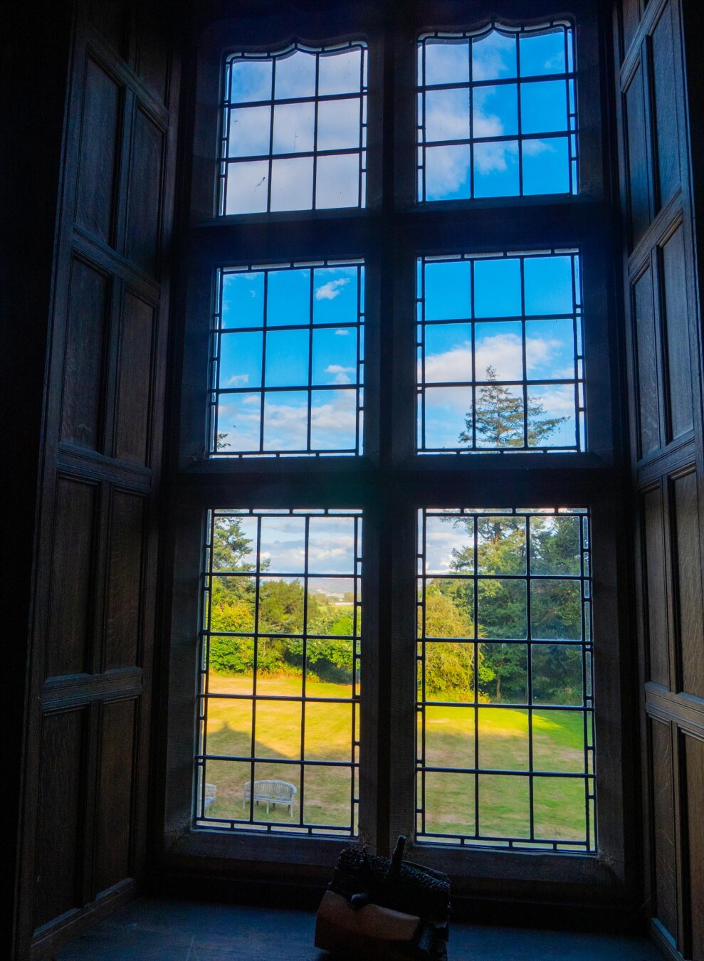 View looking out a massive, six-panel leaded glass window set in an alcove with dark wood-paneled walls. The window frames a vibrant outdoor view of a bright blue sky with fluffy white clouds, a golden grassy yard, and lush green trees.