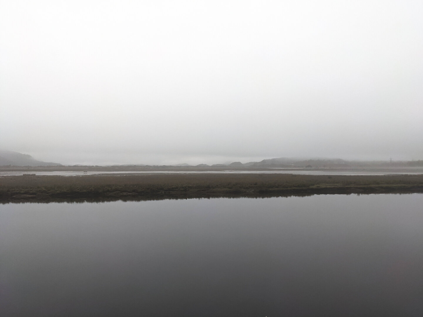 A minimalist, monochrome landscape looking over the still water of an estuary which reflects the solid overcast white/grey cloudless sky. A long, flat strip of dark marshland cuts horizontally through the center, with faint rolling hills obscured by heavy mist in the background.