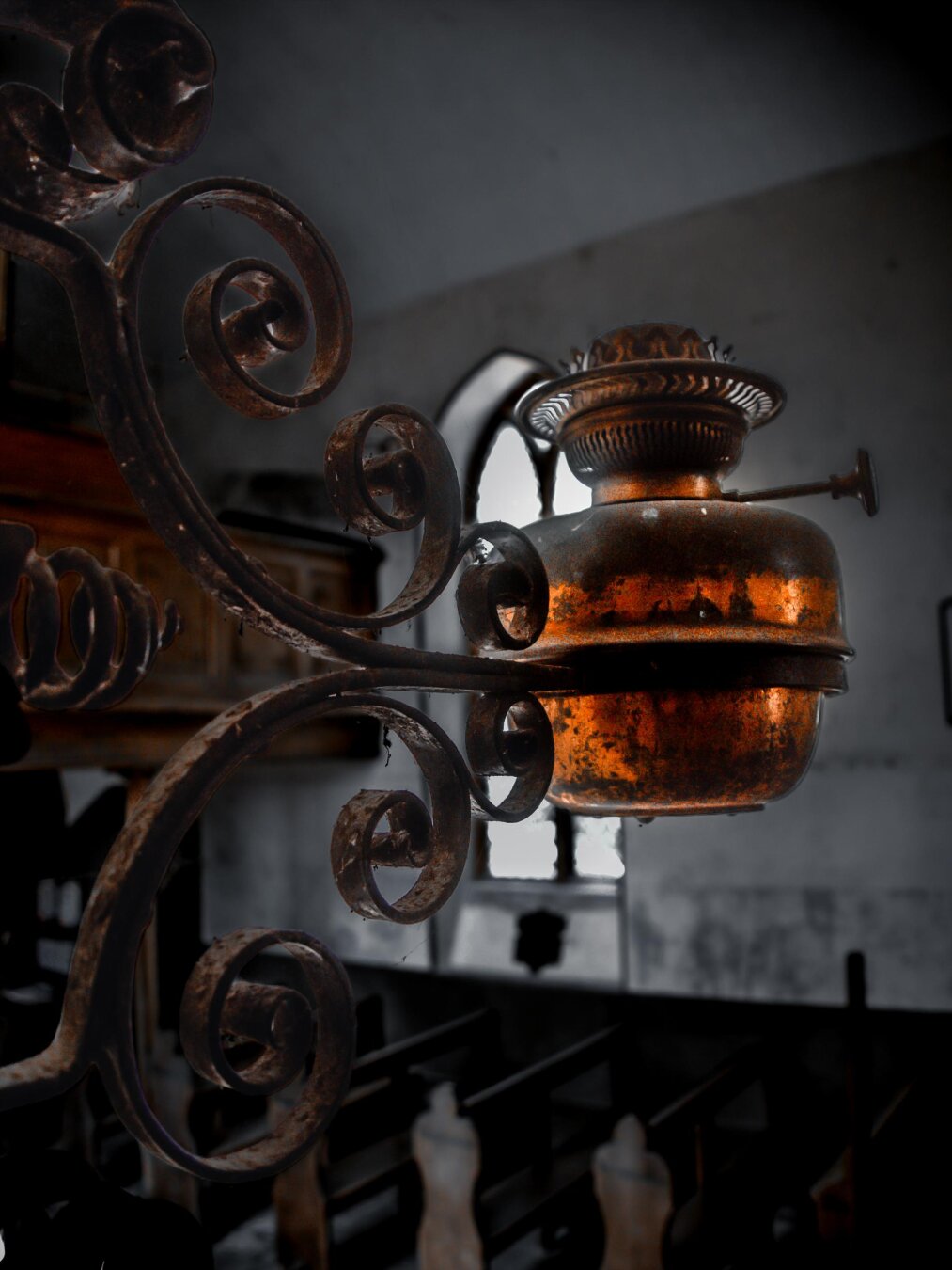 A weathered, vintage kerosene lamp burner made of aged copper or brass with an amber patina. The metallic fixture is held by thick wrought-iron details covered in light dust. In the background, rows of wooden church pews are illuminated by the light coming through an arched window.