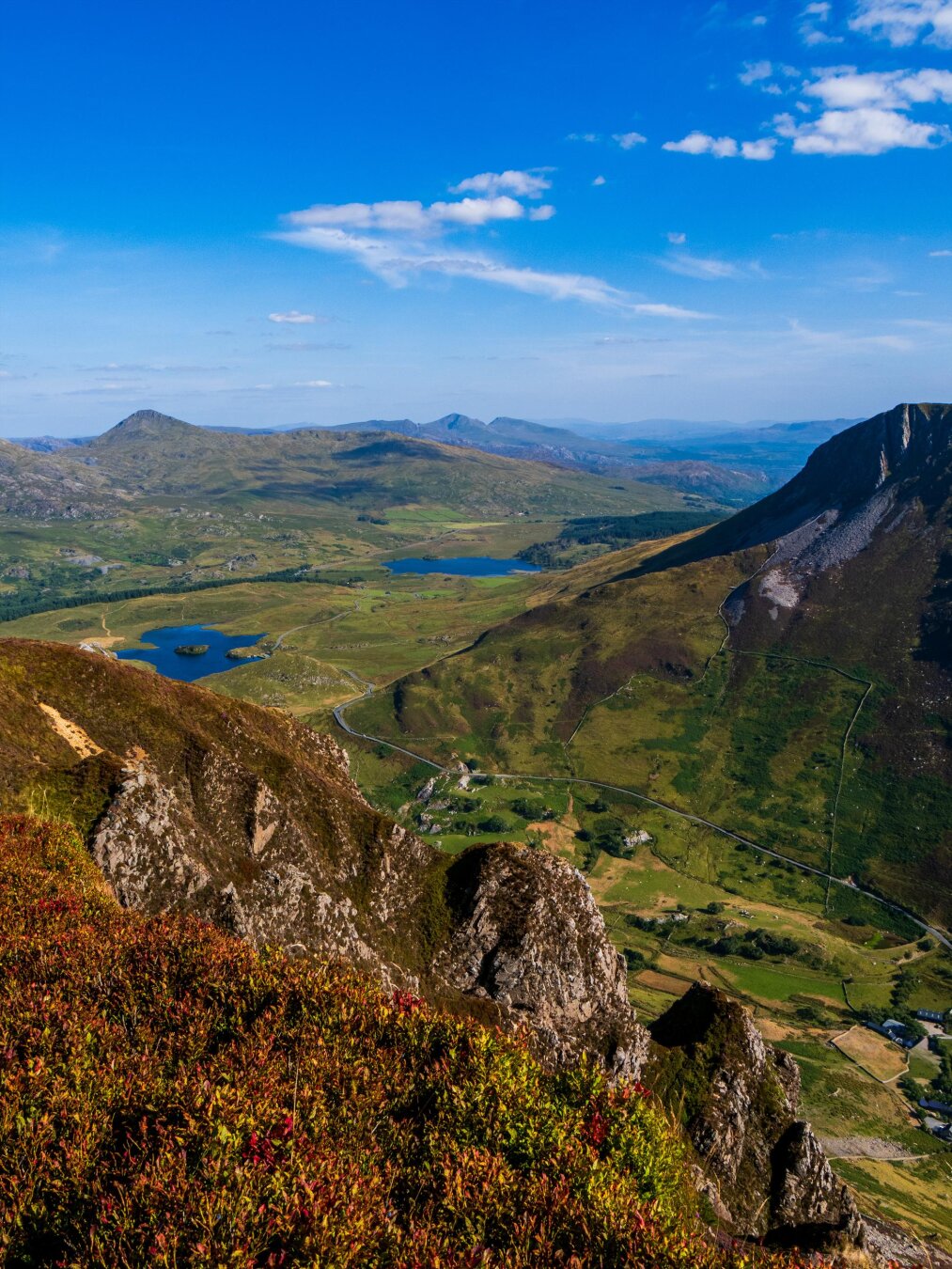 A sunny landscape from a rocky mountain ridge covered in autumn-colored heather. To the far right, a massive, steep green mountain rises over the valley below. A narrow road winds through the valley past green fields and two blue lakes. In the distance, the mountains of Eryri (Snowdonia) rise under a bright blue sky with scattered white clouds.