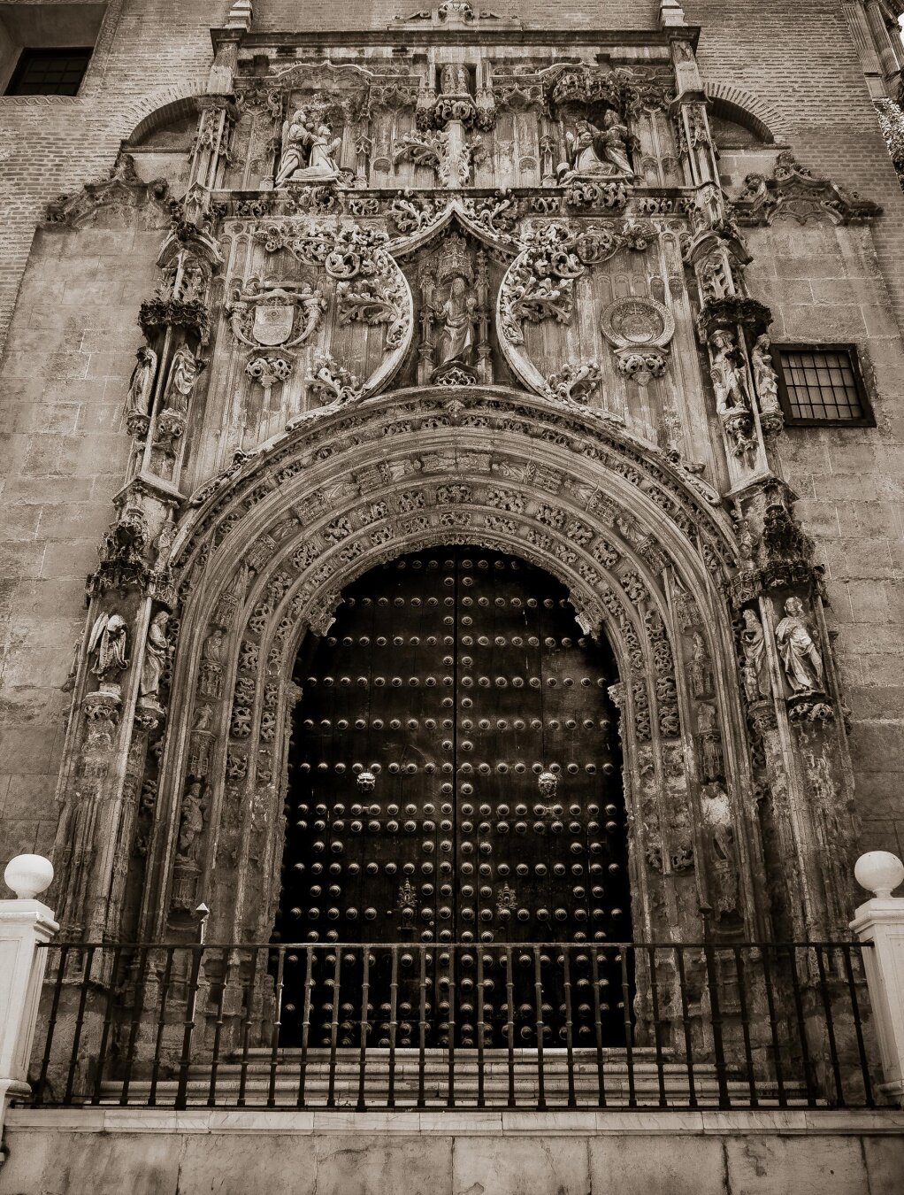 Sepia toned image looking slightly up at the 15th-century stone entrance of the Church of El Sagrario in Málaga, Spain. The large archway is framed by elaborate stonework sculpted with flowers, crests, and religious statues. A huge, imposing dark wooden double door with heavy metal studs sits in the center, visible behind a black iron fence at the bottom of the frame.