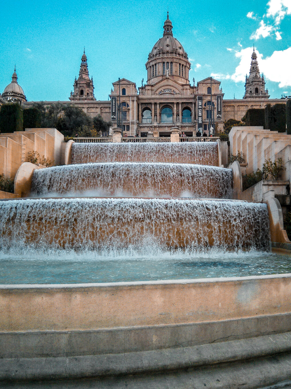 A low-angle photo of the wide, multi-level terraced waterfalls of the Montjuïc fountains in Barcelona. White water rushes over curved stone tiers into a turquoise pool. In the background, at the top of the image, the majestic Palau Nacional looms under a bright blue sky with scattered white clouds.
