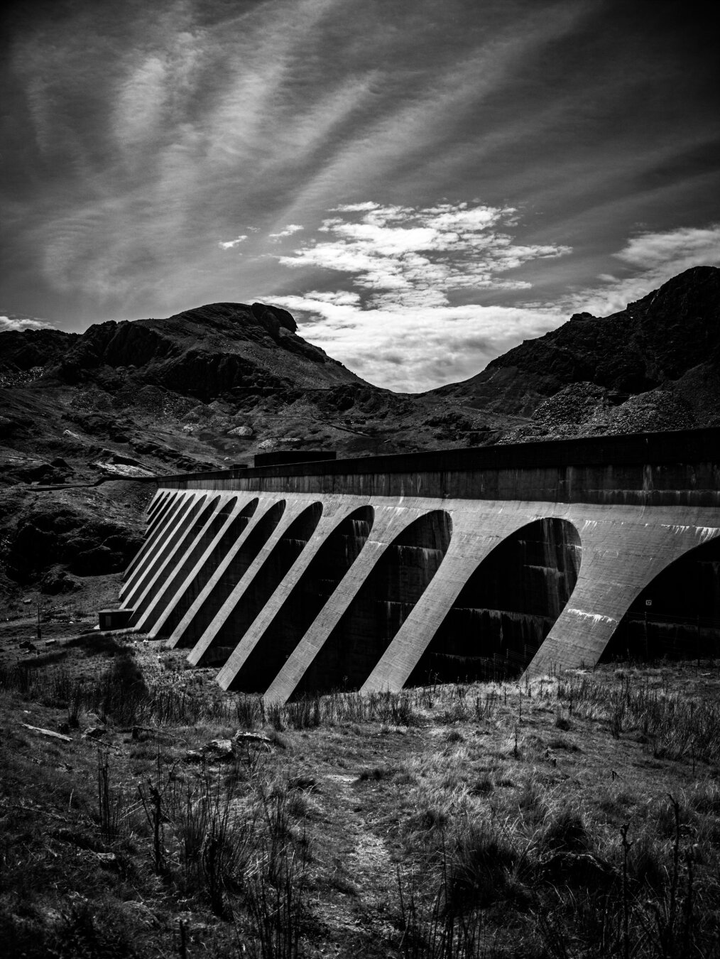 A high-contrast monochrome shot of a curved concrete dam with dark, deep arches. The dam runs across a grassy valley in Eryri (Snowdonia). In the background, dark, rocky peaks rise under a moody sky with wispy clouds.