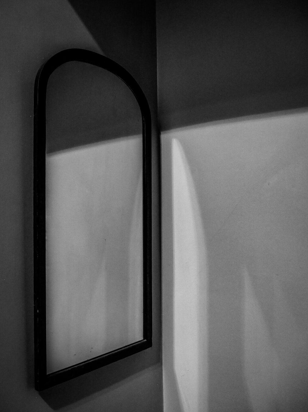 A minimalist, high-contrast monochrome image of an arched wall mirror with a thin, dark frame mounted on one wall of a bare corner. Bright lights cast angular shadows across the wall which the mirror reflects.