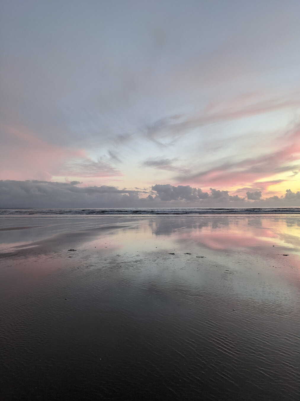 A wide, empty sandy beach at sunset, with wispy pastel pink and purple clouds streaking across a sky that turns from baby blue to golden yellow at the horizon. The sky is reflected perfectly onto the wet sand.
