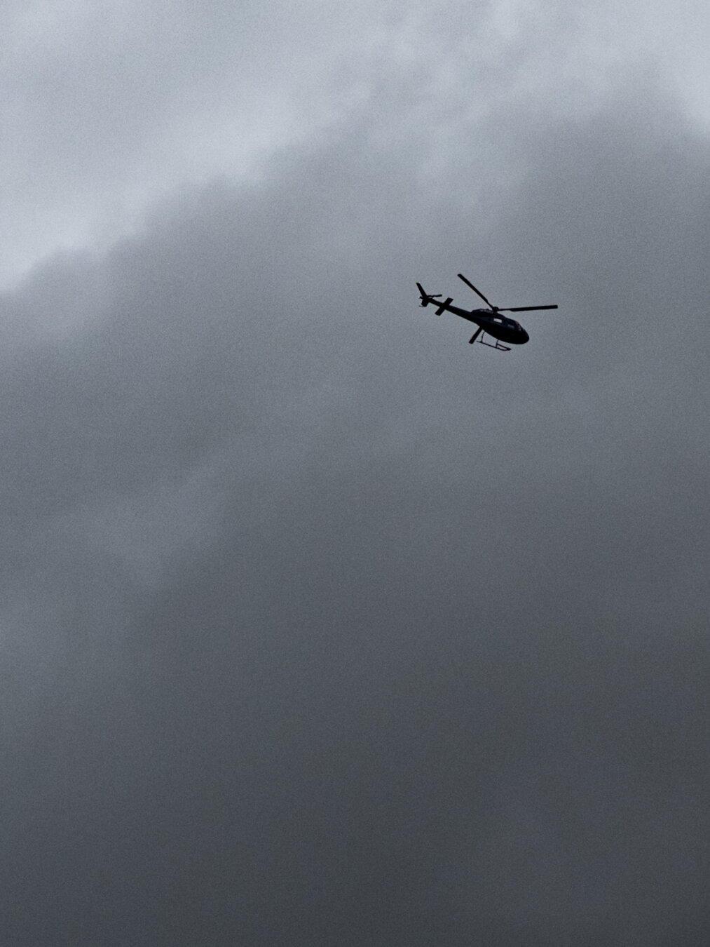 A helicopter in silhouette flying overhead in a grey, overcast sky.