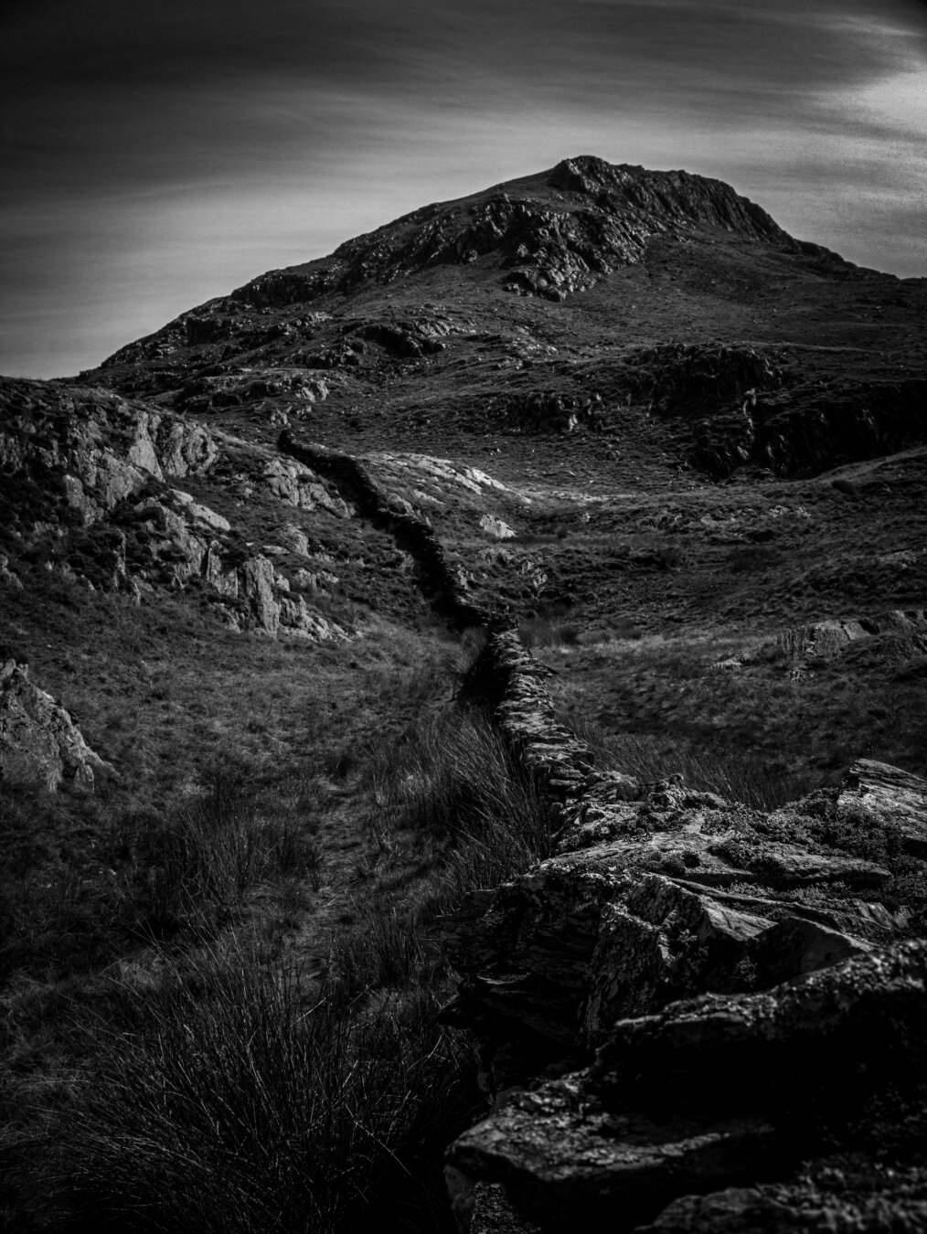 High-contrast monochrome photo of a traditional dry stone wall in Wales. The wall winds up a grassy and rocky hill towards the craggy peak of Yr Aran mountain in Eryri (Snowdonia).