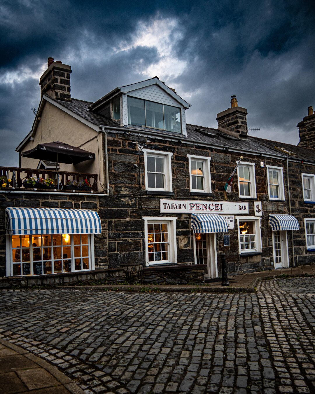A shot of a historic Welsh pub on a cobblestone street. The dark, weathered stone building features bright white window frames and matching blue-and-white striped fabric awnings over ground-floor doors and windows. In the foreground is a curved, grey cobblestone street, while overhead sky is overcast with heavy, moody clouds.