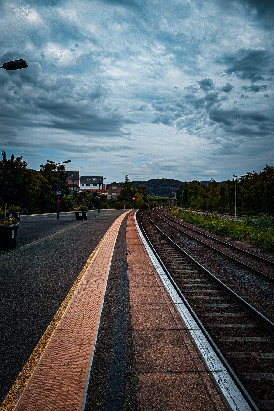 A long, empty train platform curves gently alongside multiple railway tracks under a dramatic, cloudy blue-gray sky. An orange paving strip and white edging run along the platform parallel to the tracks. To the left, several large dark planters and tall streetlights stand on the platform. Green trees and houses line the tracks ahead, with a red signal light visible in the distance where the rails curve out of view. The tracks, to the right of the image, lead toward distant hills and mountains in the background.