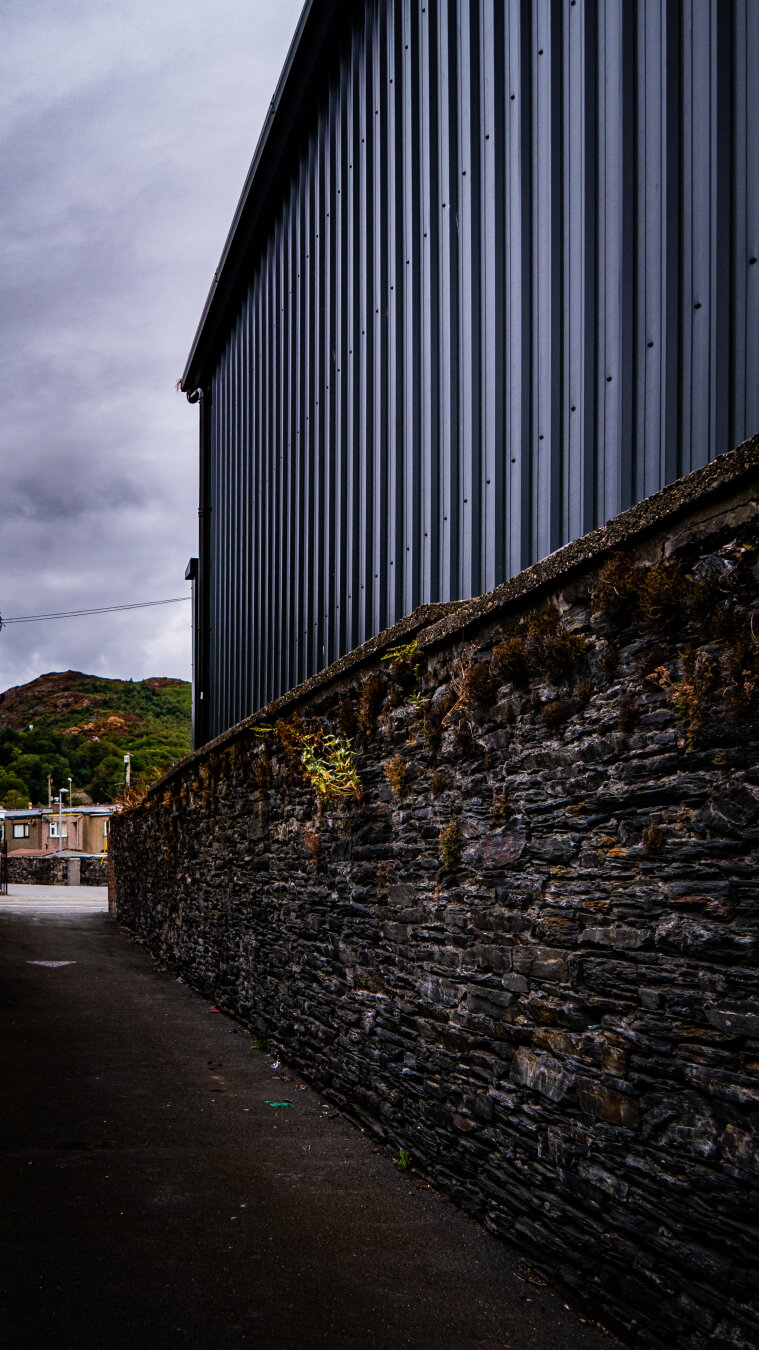 View down a narrow alleyway bordered by a heavily textured dry-stone wall behind which a towering industrial building with vertical metal cladding rises. There are weeds in greens and dry browns growing out of the stone wall. At the end of the alleyway, terraced houses can be seen just over another dry-stone wall. In the distance, a large hill rises into a moody, colourless, overcast sky.