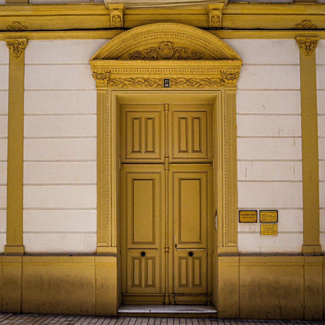 A tall, double door painted in a bold and vivid mustard yellow, surrounded by a cream-colored stone facade. There are vertical pilasters on either side of the door. Above the door is an intricately carved arched pediment. To the right of the entrance, small brass plaques are on the wall.