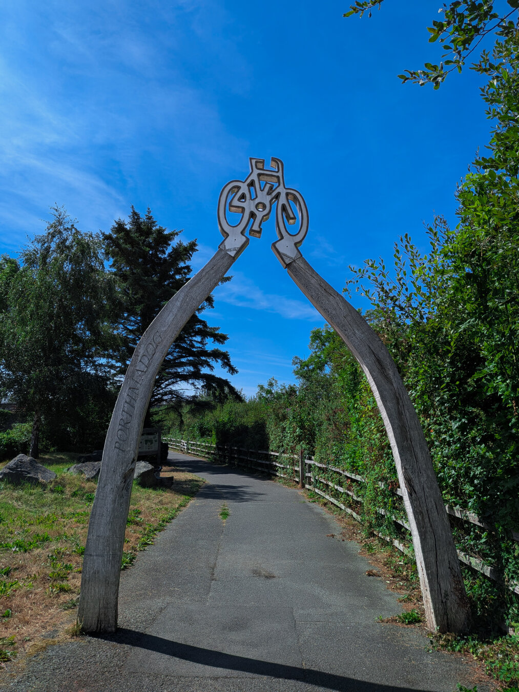 A large wooden arch carved from two massive timbers, with an integrated bicycle sculpture at the top and the word "Porthmadog" engraved on the left side. It stands over a paved pedestrian and cycling path, bordered by trees and grass under a clear blue sky.