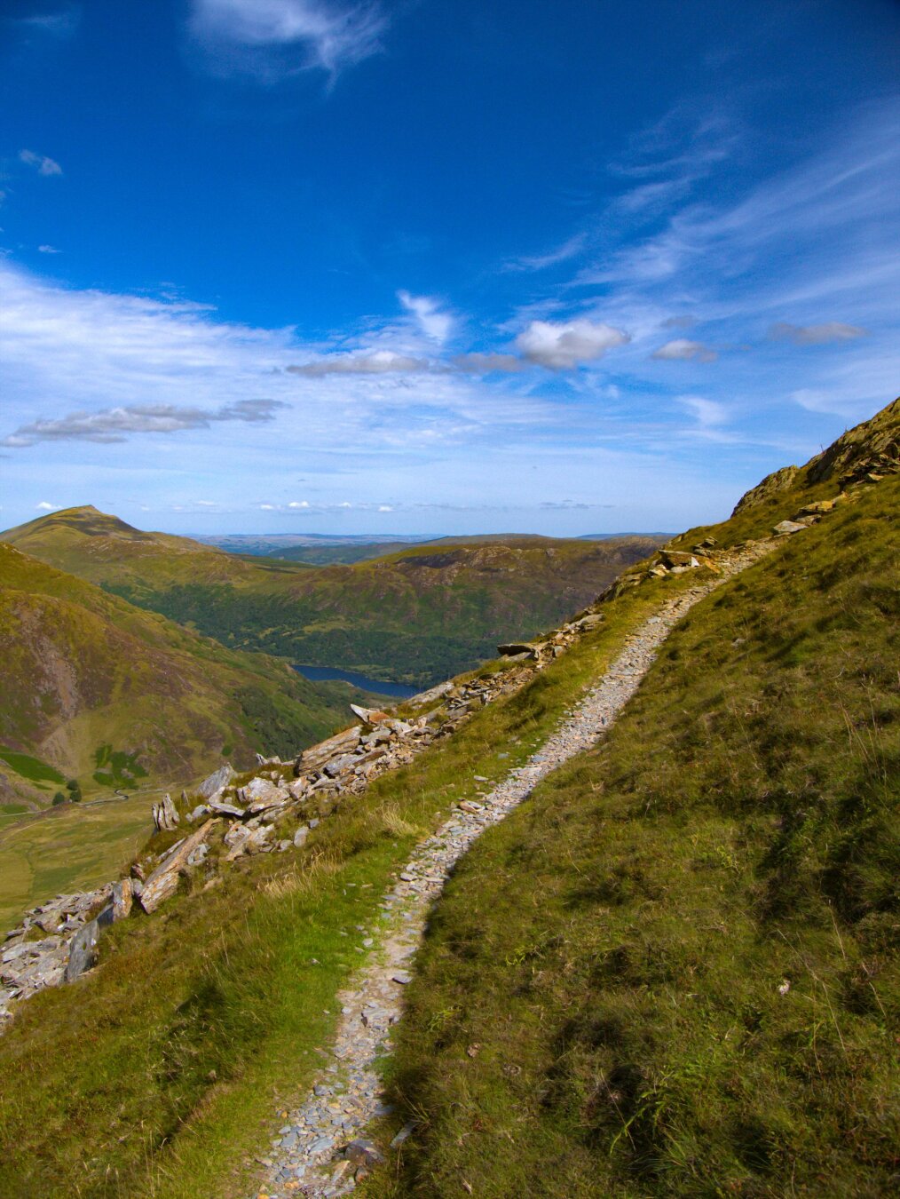 A stone and dirt footpath snakes up a steep, grassy slope of Yr Aran mountain in Eryri (Snowdonia). In the background, a blue mountain lake sits in a valley surrounded by rolling hills and lush green and brown moutains which rise into a blue sky with wispy white clouds on a sunny day.