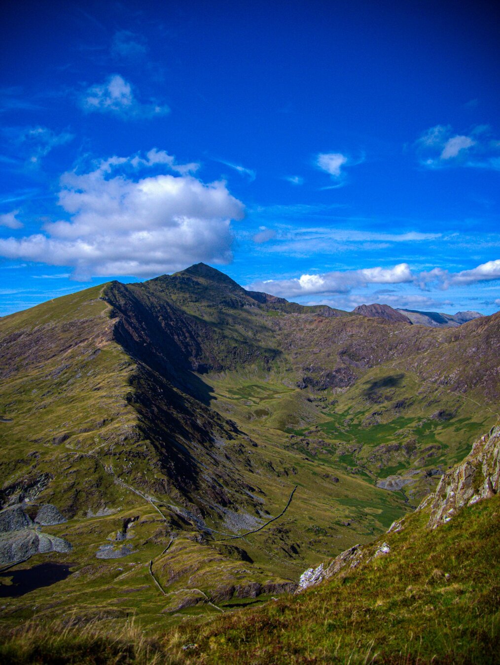 A high-angle shot looking across a grassy valley toward the pointed peak of Yr Wyddfa (Snowdon). A prominent ridge, lit up green on the left and in deep shadow on the right, climbs from the left foreground toward the summit. The valley below shows deep green patches and the faint lines of stone walls far below. A dark mountain lake sits to left of the foot of the ridge.