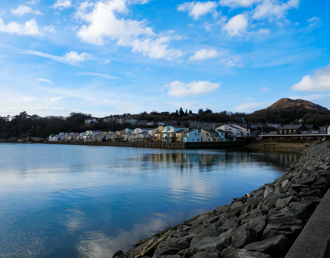 A row of pastel-colored waterfront townhouses in coral pink, light yellows, and powder blue sit overlooking the calm sea which reflects the blue sky above. In the foreground, the large gray rocks of a breakwater curve along the right edge. In the background, a large green and brown hill rises.
