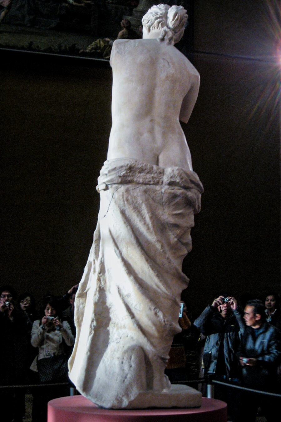 A view of the back of the Venus de Milo statue on a red circular pedestal at the Louvre Museum. The tall marble statue is nude from the waist up with a heavy cloth slipping from her hips and exposing a slight bit of tush. In the background, a crowd of tourists with cameras are standing facing her front side.